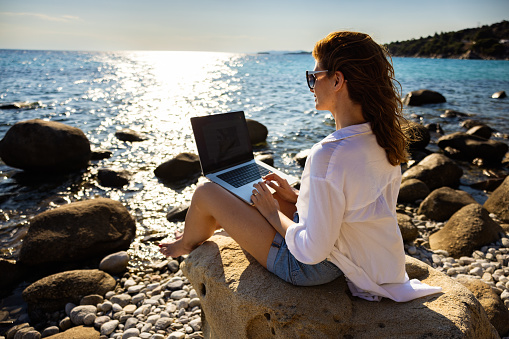 Modern young Caucasian female freelancer, working on laptop, from the rocky beach , during her vacation