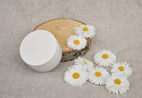 A white plastic jar for cosmetics stands on a natural wood podium and white daisy flowers for cosmetic products presentation top view on the background of linen fabric . Natural cosmetic background.