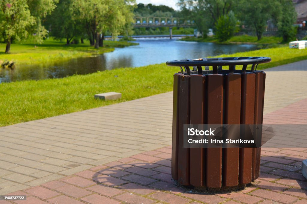 Brown wooden trash can on the street on the riverbank in the park. The dumpster is outside. Brown wooden trash can on the street on the riverbank in the park. Garbage container outside. Wastepaper Basket Stock Photo