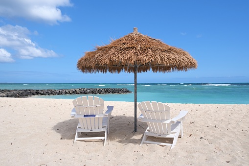 View of Diamond Head across the ocean from Kapalina Beach.  Thatched roof umbrellas and chairs on the white sand beach close to the ocean.