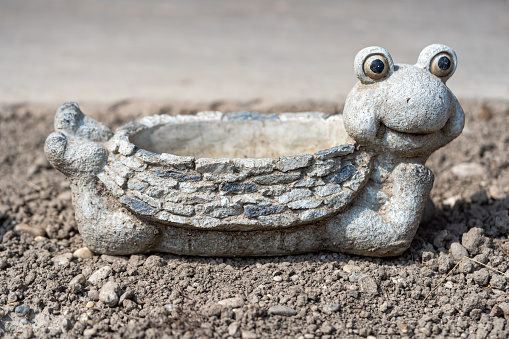Lovely frog pot made of stone in the garden on a sunny day, concrete garden decoration, hoed soil, copy space, rural concept