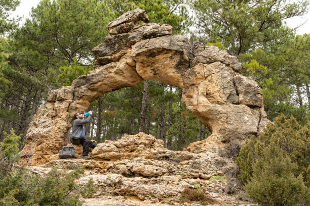 istock Young man is hydrating during a trekking while is sitting on a natural stone arch located in the middle of a pine tree forest. 1408744064