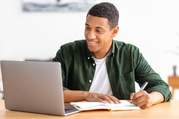 istock Smiling male student or freelancer studying remotely from home. African American male using laptop for online education, taking notes in a notebook. Online lesson, e-learning at home concept 1408746714