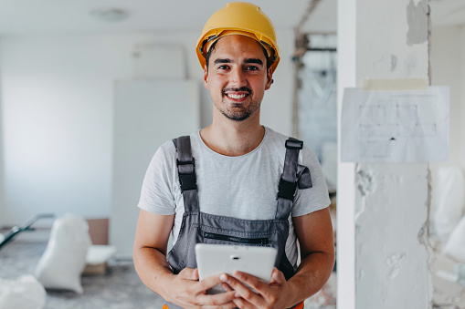 A young Caucasian male construction worker is holding a tablet and looking at the camera with a smile on his face.