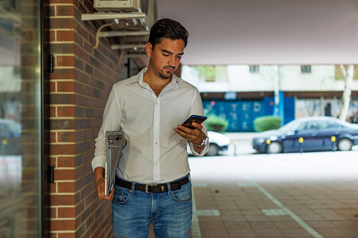 A young Caucasian businessman is standing outside of a building and typing a message on his phone.
