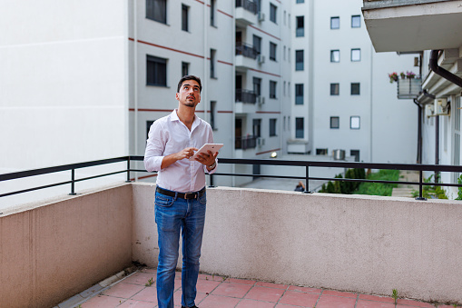 A young Caucasian businessman is standing on a balcony and looking up, while holding a tablet in his hands.