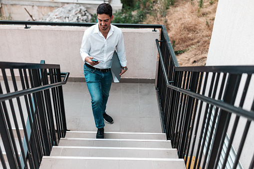 A young Caucasian businessman is walking up a flight of stairs with a warm smile on his face, while holding a phone and a file folder.