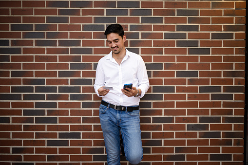 A young Caucasian businessman is leaning against a brick wall, while making an online purchase with a smile on his face.