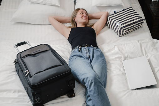 Woman with suitcase on bed in hotel having rest while traveling. Relaxed happy Traveler with bags in hostel Top view. Enjoying trip booking apartments. Arriving at resort