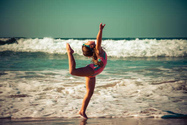 Young girl in bathing suit dances on the beach with ocean waves behind her