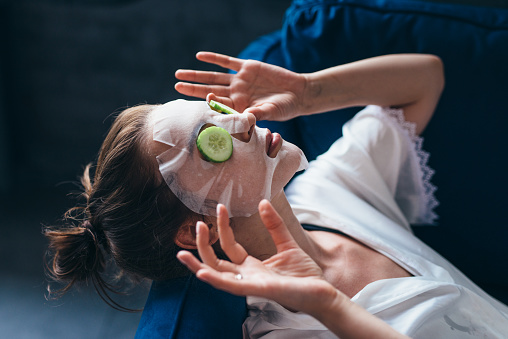 Woman lies on a couch with a sheet mask over her face and cucumber slices.