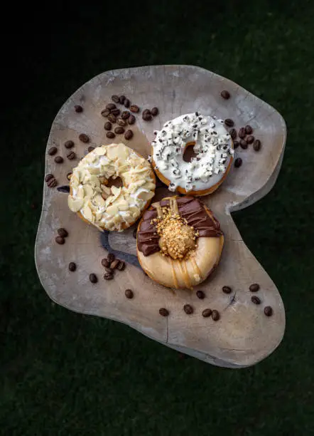 Three doughnuts covered with Cookies and creme, Almendras and Choco peanut butter on wooden cutting board in dark background. The concept of delicious glazed donuts, Top view, Space for text, Selective Focus.