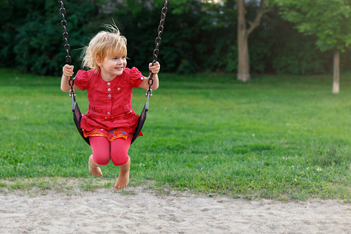 Happy smiling child swinging in park. Kid at playground. Little girl in red dress sitting on swing.