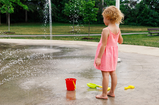 Little child playing with water and toys at splash pad in the local public park during hot summer day. Small beautiful girl in pink dress having fun at fountain playground