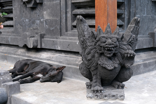 Dog sleeping by an ancient statue at Pura Goa Lawah, a Hindu temple in Klungkung, Bali, Indonesia. Pura Goa Lawah is noted for built around a cave opening which is inhabited by bats, hence its name, the Goa Lawah or \