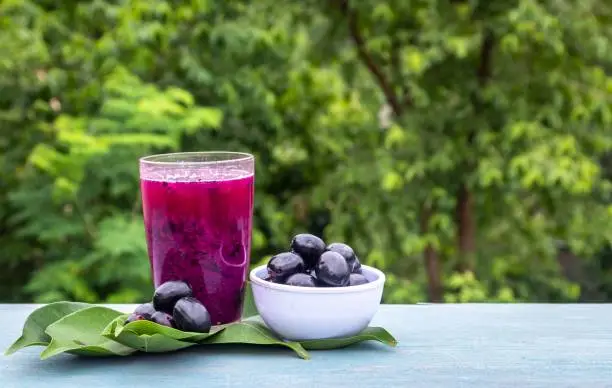 Jamun or Black Plum Juice in a Glass with Fruit in a Bowl with Leaves Isolated on Wooden  Background with Copy Space for Texts Writing, Also Known as Malabar Plum, Syzygium Cumini or Jambolan in Horizontal Orientation.