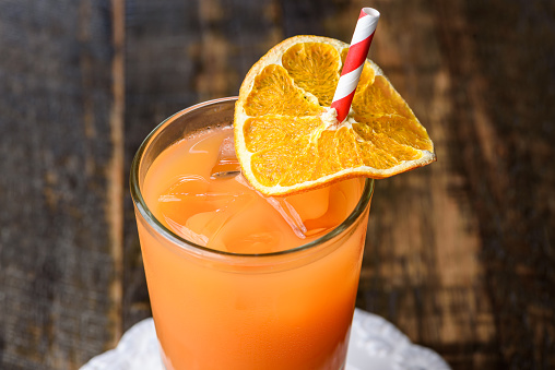 An orange and carrot juice in a tall glass, on a white plate and against a wooden tabletop background