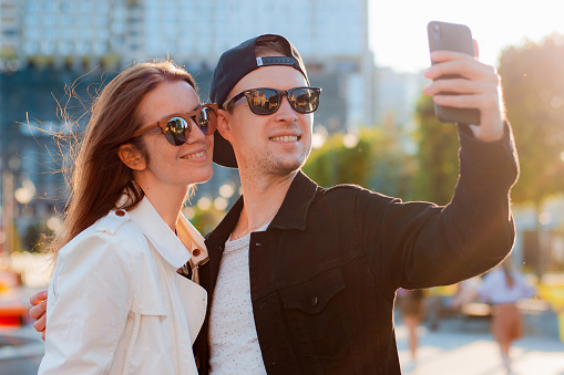 Fashionable couple taking selfie by mobile phone. man and a woman in sunglasses meeting friends in the city date photo on a smartphone against the sunset.