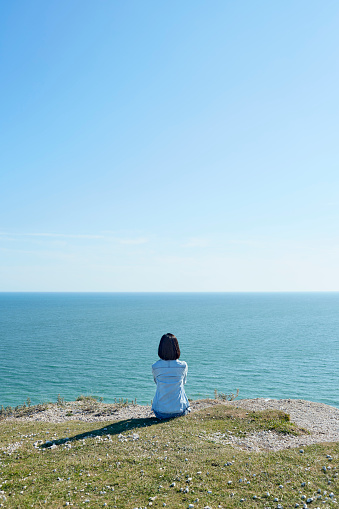 A Hong Kong Chinese Asian woman sitting by the sea, beneath a clear sunny blue sky.