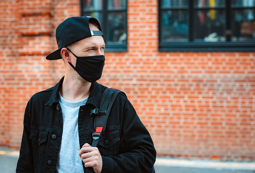 young fashionable man youth casual wear protective black mask walks through urban city sunset. trendy teen guy walks around city coronavirus pandemic. Spring. Modern Fashion 2020.