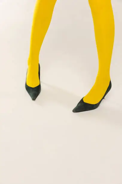 Cropped image of female legs in yellow bright tights and heels isolated over grey studio background. Colorful outfit. Concept of retro fashion, art photography, style, queer, beauty