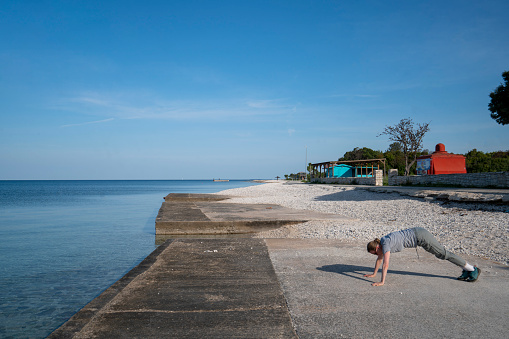 Side view of senior woman exercising push-ups 
on the beach in spring morning. Shi is in small place Peroj near Pula in Istria, Croatia with beach facilities in background.