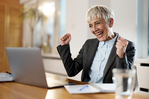Cheerful mature businesswoman celebrating her success after receiving good news over laptop at home office.