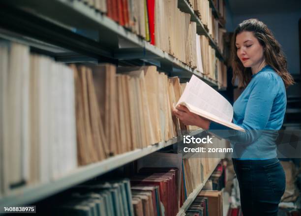 Female Postgrad Standing By The Shelf In The Library And Going Through A Book Stock Photo - Download Image Now