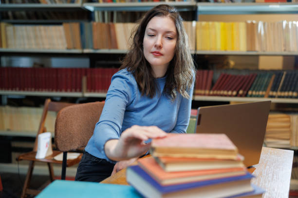 istock Female post-grad using laptop at the desk in the library and taking book from stack 1408839017