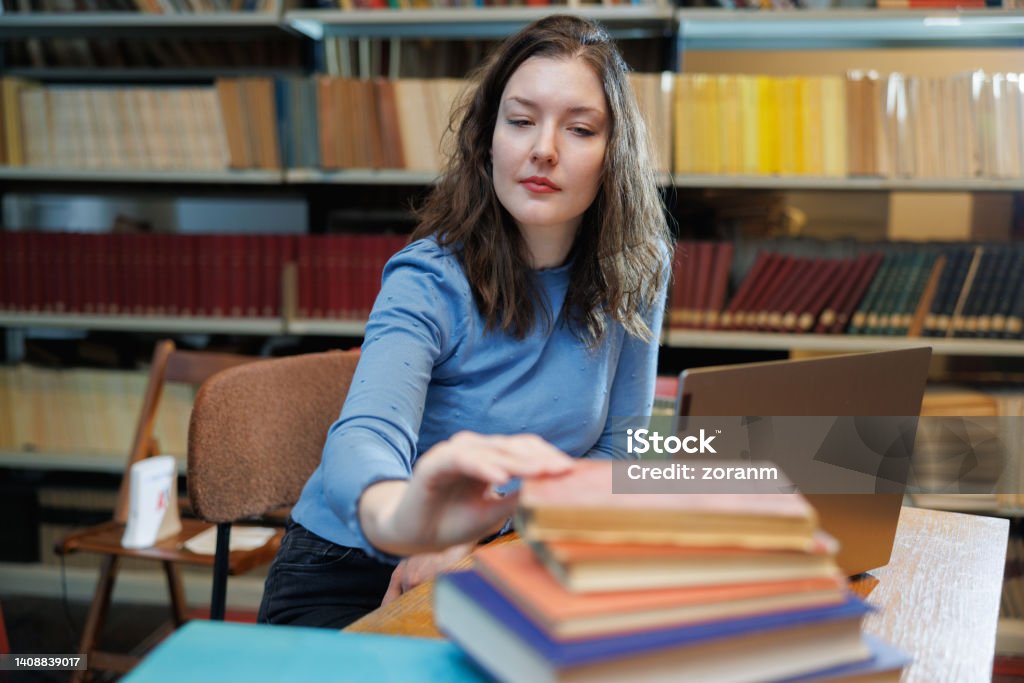 Female post-grad using laptop at the desk in the library and taking book from stack Beautiful university student sitting at the desk in the library with laptop, picking up hardcover book from stack 30-34 Years Stock Photo