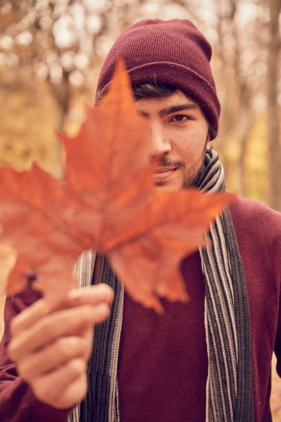 White Caucasian man looking at camera clutching a brown five-pointed leaf covering one eye in a park in autumn