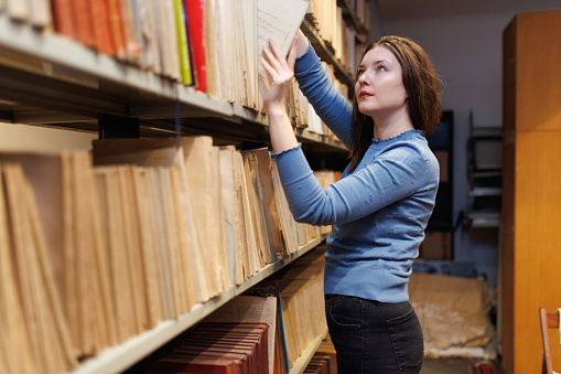 Beautiful woman working at the university library, standing by the shelves and archiving material