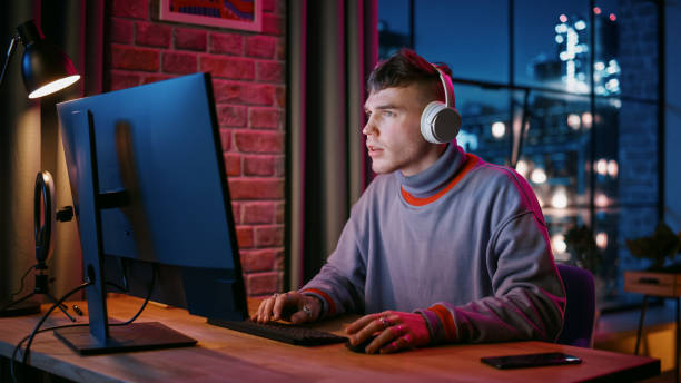 istock Young Handsome Man in Headphones Using Computer in Stylish Loft Apartment in the Evening. Creative Male Smiling, Working from Home, Browsing Videos on Social Media. Urban City View from Big Window. 1408840557
