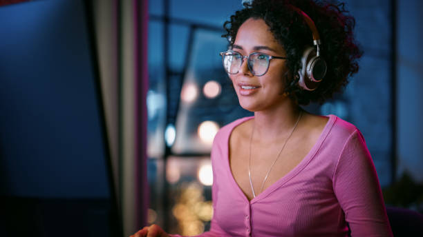 istock Young Attractive Black Woman Using Computer in Stylish Loft Apartment in the Evening. Creative Female Smiling, Checking Social Media, Browsing Videos on Internet. Urban City View from Big Window. 1408840869