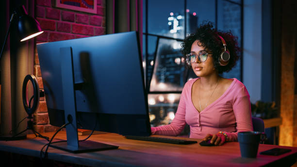 istock Young Attractive Black Woman Using Computer in Stylish Loft Apartment in the Evening. Creative Female Smiling, Checking Social Media, Browsing Videos on Internet. Urban City View from Big Window. 1408841432
