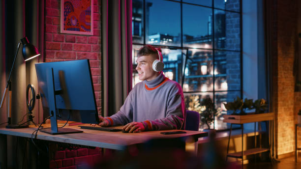 istock Young Handsome Man in Headphones Using Computer in Stylish Loft Apartment at Night. Creative Male Working from Home, Browsing Internet and Social Media. Urban City View from Big Window. 1408841833