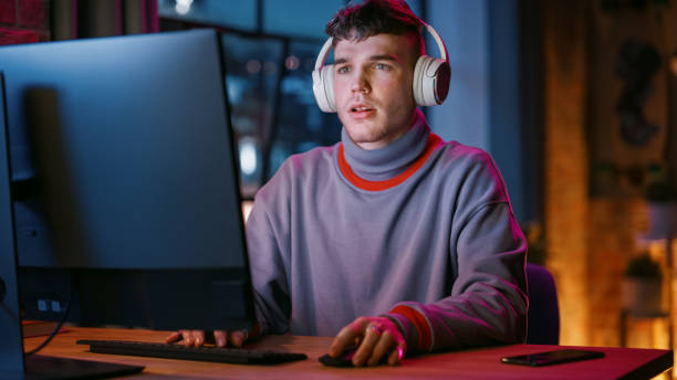 istock Young Handsome Man in Headphones Using Computer in Stylish Loft Apartment in the Evening. Creative Male Smiling, Working from Home, Browsing Videos on Social Media. Urban City View from Big Window. 1408841948