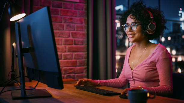 istock Young Attractive Black Woman Using Computer in Stylish Loft Apartment in the Evening. Creative Female Smiling, Checking Social Media, Browsing Videos on Internet. Urban City View from Big Window. 1408841979