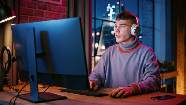 istock Young Handsome Man in Headphones Using Computer in Stylish Loft Apartment in the Evening. Creative Male Smiling, Working from Home, Browsing Videos on Social Media. Urban City View from Big Window. 1408842035