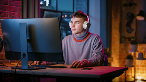 istock Young Handsome Man in Headphones Using Computer in Stylish Loft Apartment in the Evening. Creative Male Smiling, Working from Home, Browsing Videos on Social Media. Urban City View from Big Window. 1408842068