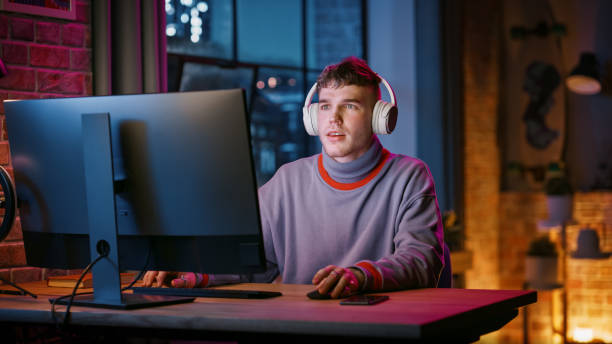 istock Young Handsome Man in Headphones Using Computer in Stylish Loft Apartment in the Evening. Creative Male Smiling, Working from Home, Browsing Videos on Social Media. Urban City View from Big Window. 1408842079