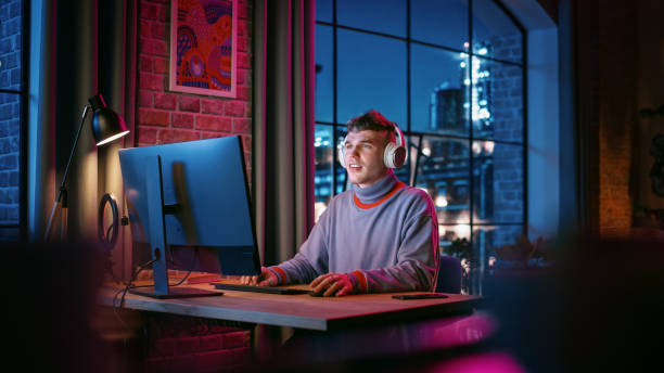 istock Young Handsome Man in Headphones Using Computer in Stylish Loft Apartment at Night. Creative Male Working from Home, Browsing Internet and Social Media. Urban City View from Big Window. 1408842111