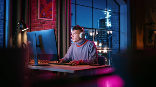 junger gutaussehender mann mit kopfhörern und computer in stilvoller loft-wohnung am abend. kreative männliche lächeln, arbeiten von zu hause aus, durchsuchen von videos in sozialen medien. urbaner blick auf die stadt aus dem großen fenster. - loft apartment design studio office studio stock-fotos und bilder