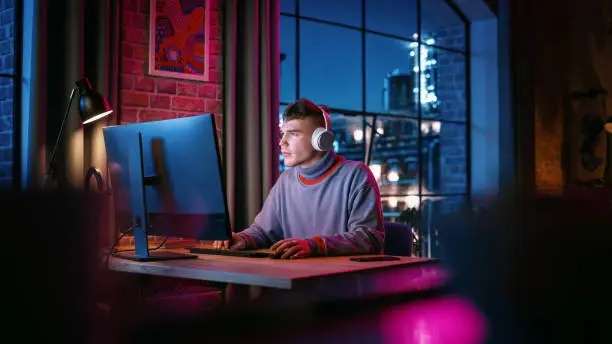Young Handsome Man in Headphones Using Computer in Stylish Loft Apartment in the Evening. Creative Male Smiling, Working from Home, Browsing Videos on Social Media. Urban City View from Big Window.