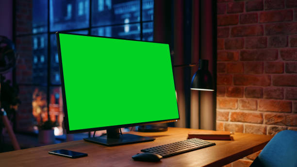 istock Desktop Computer Monitor Standing on a Table with a Green Screen Chromakey Mock Up Display. Cozy Empty Loft Apartment with Big Window with a Lamp, Notebook and Smartphone on the Table. 1408842262
