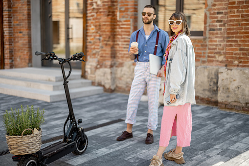 Young stylish colleagues meet together standing with electric scooter near office outdoors. Modern hipsters during coffee break, sustainable lifestyle concept