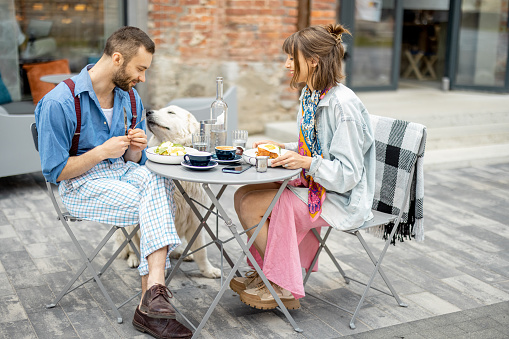 Stylish man and woman have delicious breakfast, sitting with cute dog at cafe terrace. Friends spending time together during a breakfast