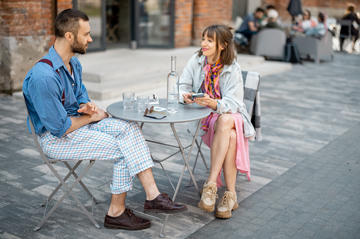 Stylish couple of colleagues have a conversation while sitting at cafe terrace in modern office district. Couple spending time together drinking water by the table outdoors