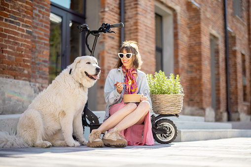 Young stylish woman has a coffee break, eating some take away food while sitting with her dog on electric scooter near office outdoors. Modern and sustainable lifestyle concept
