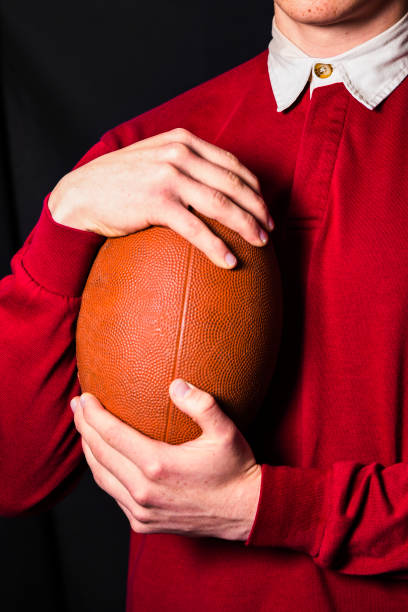 joven jugador de rugby con camiseta deportiva retro roja sosteniendo pelota de rugby en las manos sobre fondo negro - men button down shirt black smug fotografías e imágenes de stock